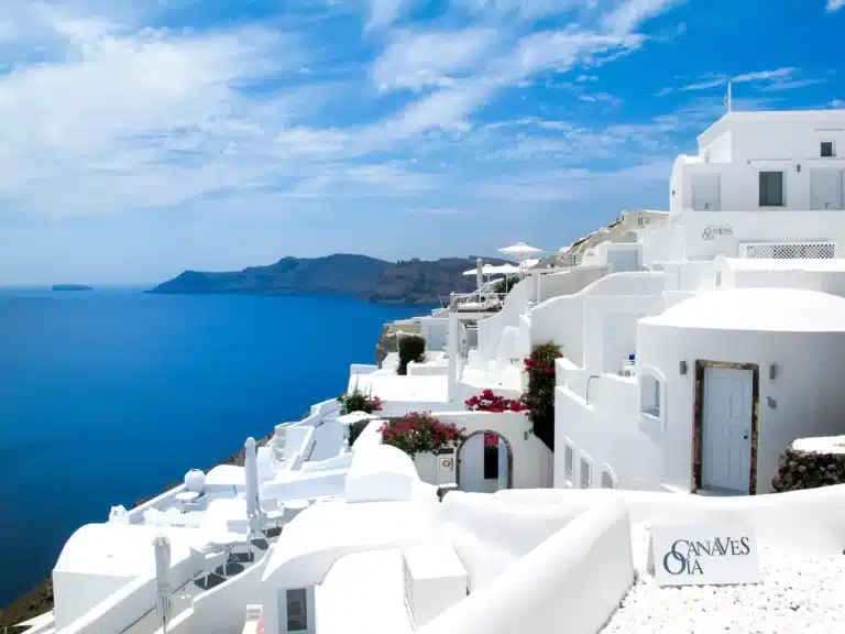 Weiß getünchte Gebäude mit blauen Akzenten entlang einer Klippe mit Blick auf den Ozean von einem Katamaran aus an einem strahlend sonnigen Tag in Santorin, Griechenland.
