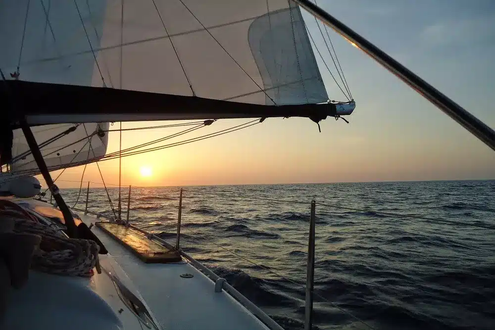 Eine Segelyacht auf See bei Sonnenuntergang, die Sonne steht tief am Horizont und wirft einen sanften goldenen Schimmer auf das Wasser. Die Segel des Bootes sind mit Wind gefüllt, was das Gefühl eines