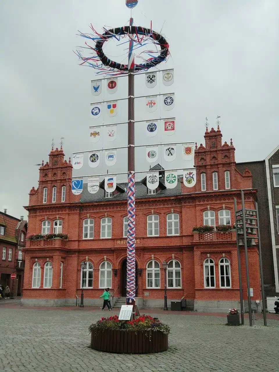 Ein Maibaum mit bunten Bändern steht auf einem Platz vor einem traditionellen roten Backsteingebäude mit verzierten Fenstern, geschmückt mit verschiedenen Wappenschildern unter einem bewölkten Himmel. In der Nähe, Enthusiasten