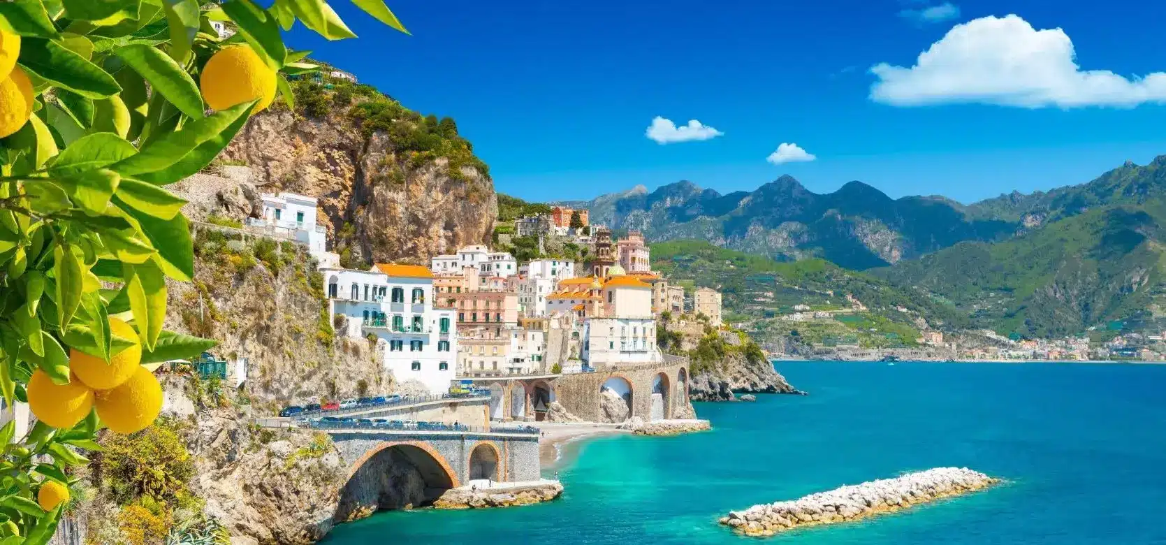 Ein malerischer Blick auf die Amalfiküste in Italien mit lebendigen, bunten Gebäuden auf Klippen mit Blick auf das blaue Meer, wo ein Katamaran sanft unter einem klaren Himmel segelt,