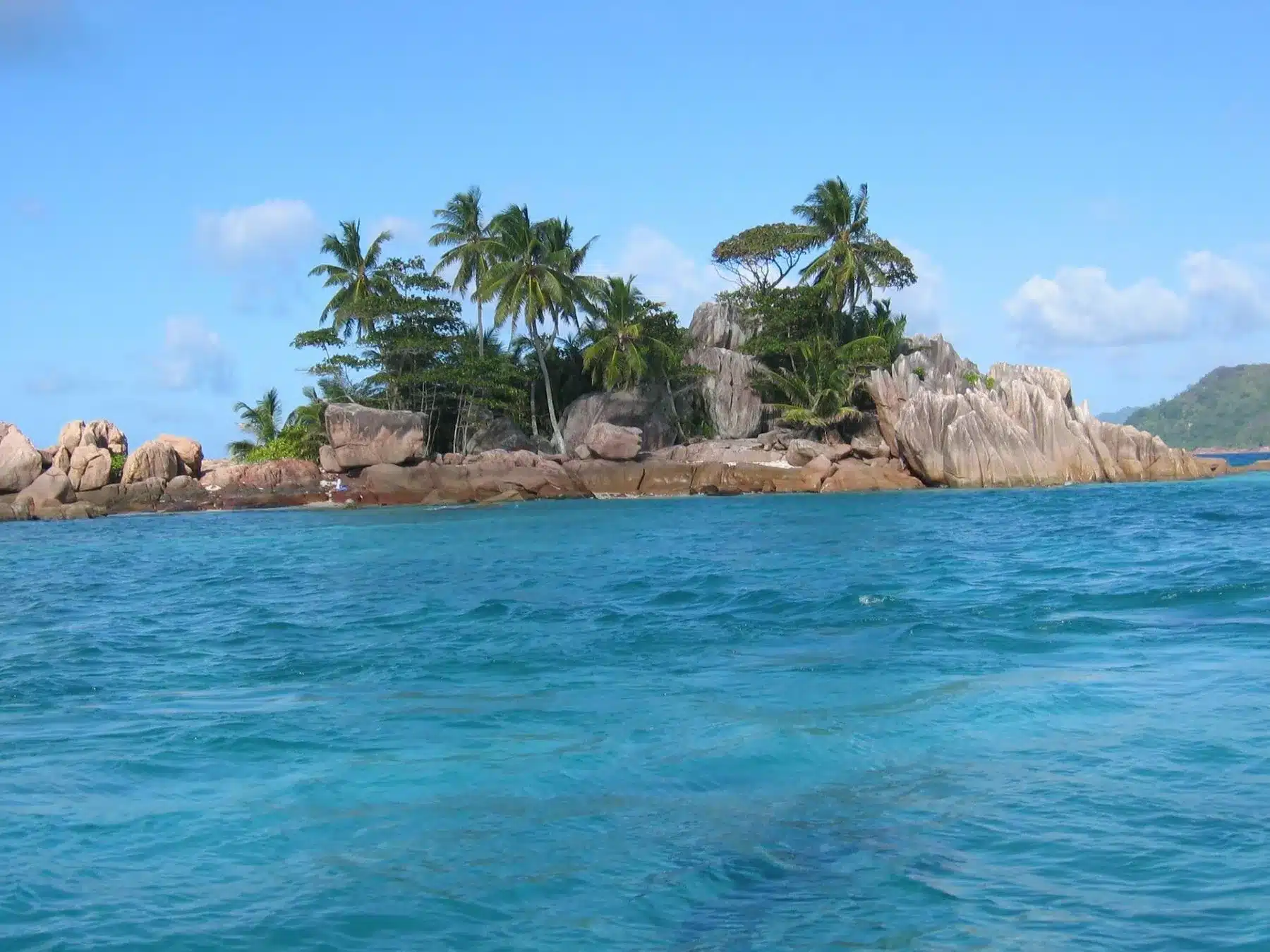 Ein malerischer Blick auf eine tropische Insel mit Felsformationen und üppigen Palmen vor einem klaren blauen Himmel, gesehen vom türkisfarbenen Ozean während eines Segelabenteuers.