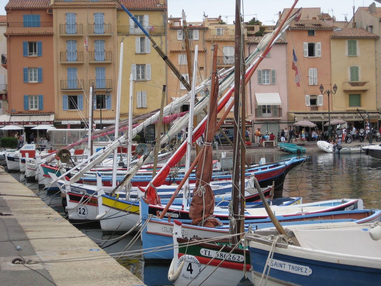 Traditionelle hölzerne Fischerboote und eine Segelyacht liegen in einem ruhigen Hafen vor Anker, im Hintergrund sind farbenfrohe Gebäude zu sehen, unter einem bewölkten Himmel. Flaggen flattern leicht im Wind.