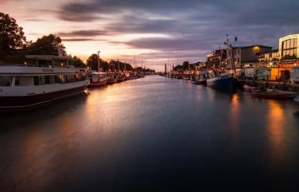 Sonnenuntergang über einem ruhigen Fluss mit Booten, die an von Bäumen gesäumten Ufern festgemacht sind, und Gebäuden, die vom schwindenden Abendlicht beleuchtet werden und sich sanft im Wasser spiegeln. Unter den Booten sticht eine Segelyacht hervor