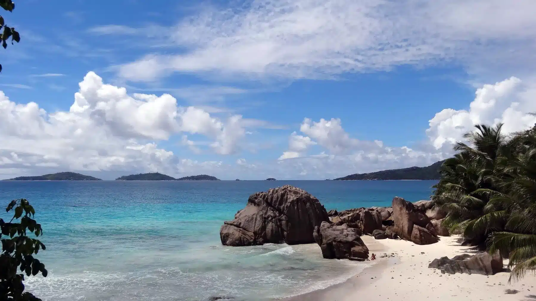 Tropische Strandszene mit klarem, türkisfarbenem Wasser, einem Sandstrand, Palmen auf der linken Seite und einer Segelyacht in der Nähe der großen Felsbrocken am Ufer unter einem teilweise bewölkten blauen Himmel.