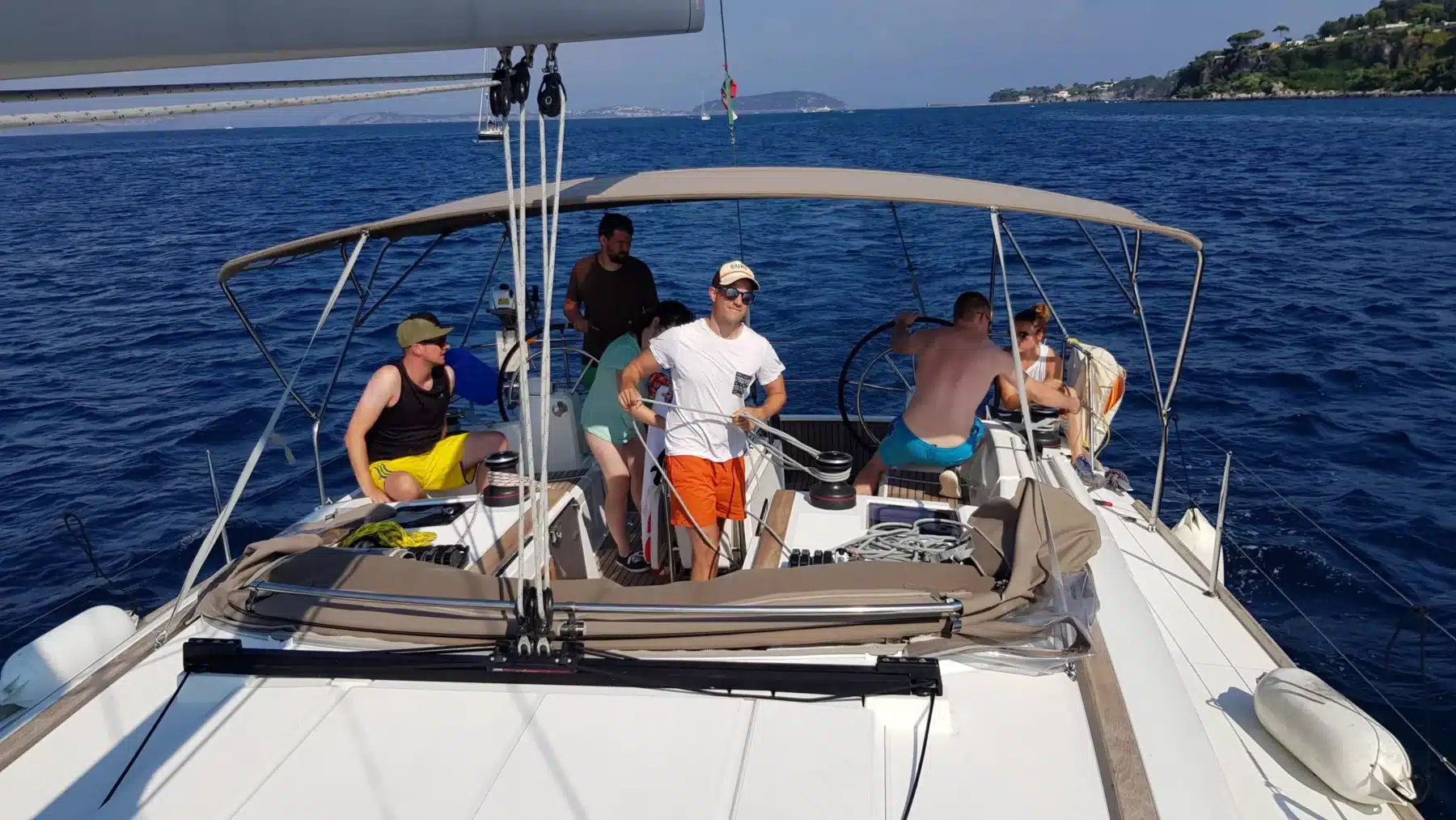 Eine Gruppe von Menschen genießt einen sonnigen Tag auf einem Katamaran. Ein Mann steht und lächelt in die Kamera, andere sitzen oder stehen um ihn herum. Im Hintergrund ist die Küste zu sehen.