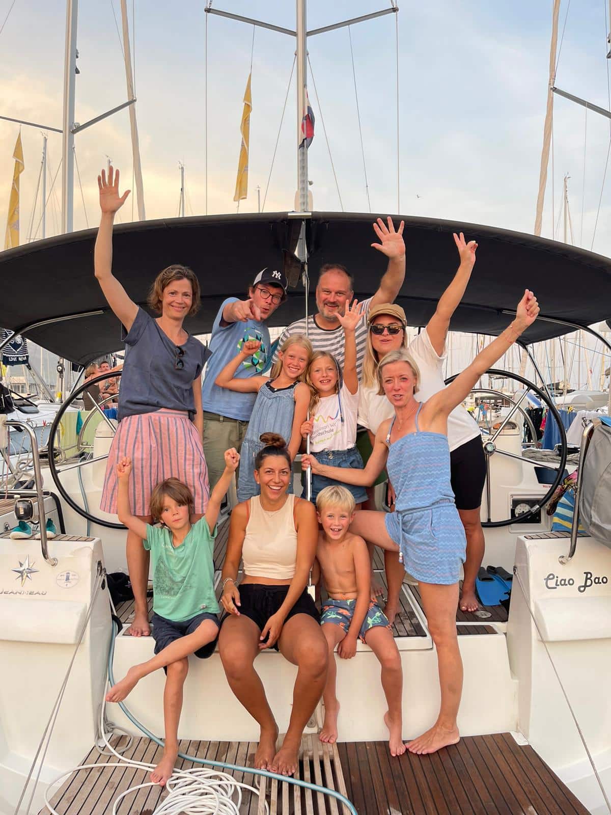 Eine Gruppe von neun Personen, darunter Erwachsene und Kinder, posiert fröhlich auf einem Katamaran, der in einem Yachthafen angedockt ist. Sie lächeln, und einige schwenken ihre Arme in der Luft. Im Hintergrund sind Masten und Takelage anderer Boote im Yachthafen zu sehen, die bereit für einen aufregenden Segeltörn sind.
