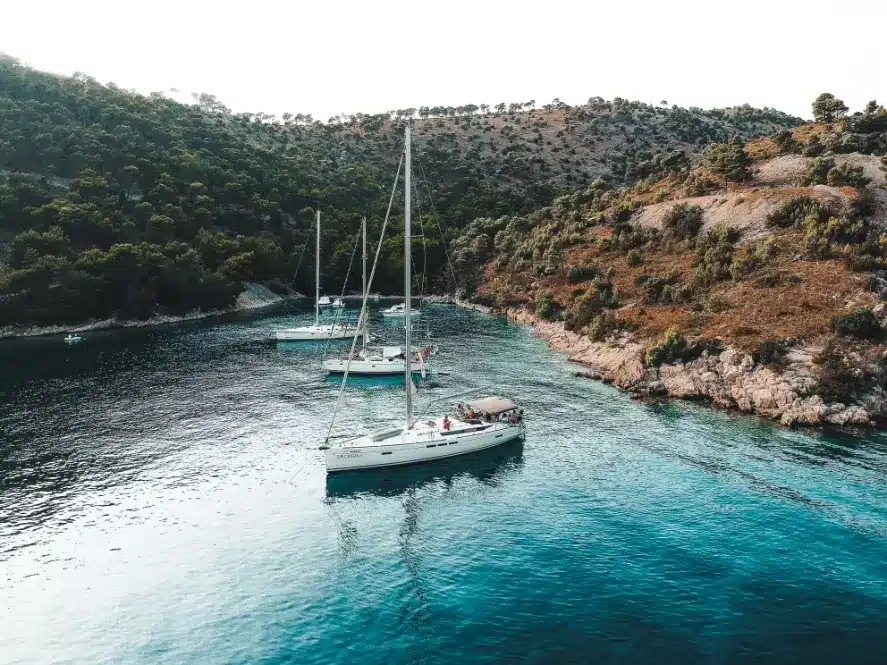 Luftaufnahme von zwei Segelbooten, darunter ein Katamaran, die in einer ruhigen Bucht mit klarem blauen Wasser vor Anker liegen, umgeben von üppigen grünen Hügeln.