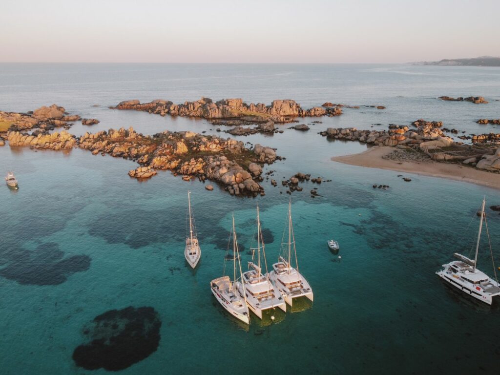 Luftaufnahme mehrerer Segelboote und Katamarane, die in klarem, türkisfarbenem Wasser nahe einer felsigen Küste vor Anker liegen. Das Wasser reflektiert das warme Licht des frühen Abends und kleine Inseln liegen verstreut im ruhigen Meer. Auf diesem idyllischen Segeltörn ist in der Ferne der Horizont sichtbar, mit sanften Wellen, die ans Ufer schlagen.