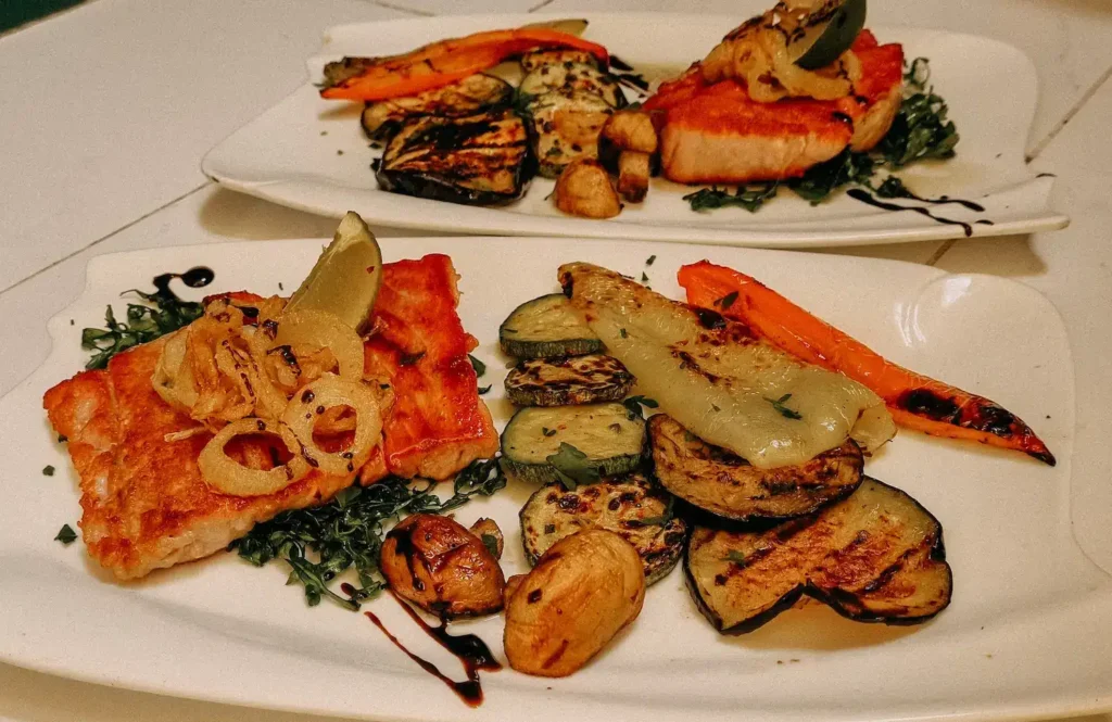 Auf zwei rechteckigen Tellern liegt jeweils eine Portion gegrillter Lachs, garniert mit sautierten Zwiebeln. Der Lachs, der an ein feines Essen an Bord eines Luxuskatamarans erinnert, wird von verschiedenen gegrillten Gemüsesorten begleitet, darunter Zucchini, Paprika, Pilze und Auberginen auf einem Bett aus Grünzeug. Bordleben und kulinarische Highlights: Ein Genuss für Gourmets