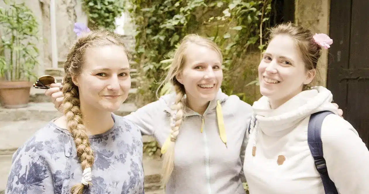 Drei Frauen lächeln und posieren zusammen auf einer Segelyacht, zwei mit geflochtenen, mit Blumen geschmückten Haaren und eine trägt einen Rucksack.