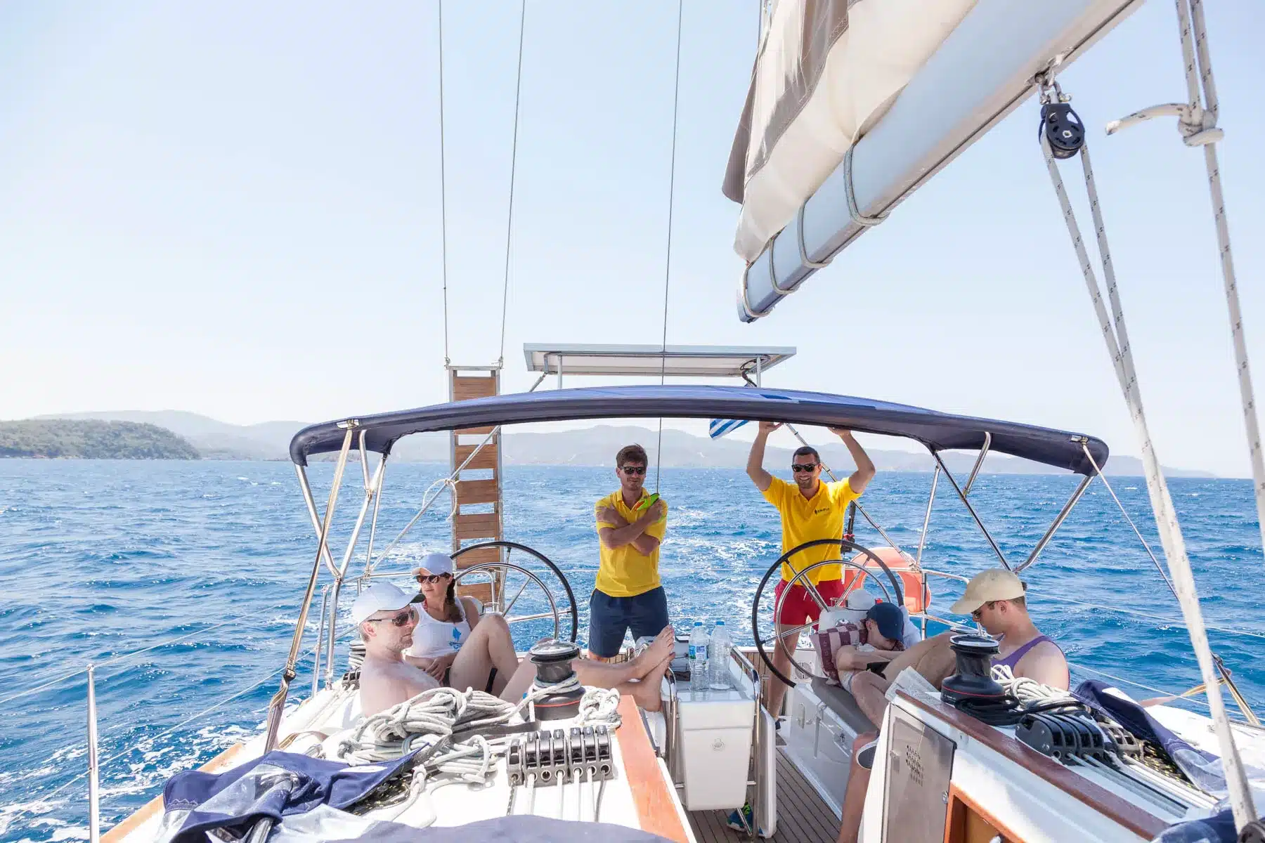 Eine Gruppe von Menschen entspannt sich auf einer Segelyacht unter klarem Himmel. Zwei Personen in gelben Hemden stehen am Steuer, während andere auf dem Deck sitzen und faulenzen. Im Hintergrund sind blaue Meereswellen und fernes Land zu sehen.