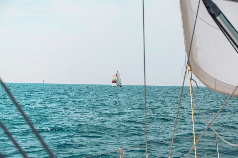 Blick von einem Katamaran mit teilweiser Sicht auf das Segel über die ruhige See auf ein anderes, in der Ferne gelegenes Segelboot mit einem bunten Segel.