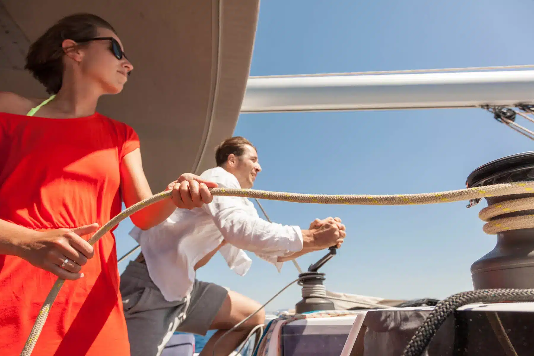 Zwei Personen auf einer Segelyacht; eine Frau in einem roten Kleid beobachtet, wie ein Mann in einem weißen Hemd und einer schwarzen Hose unter einem klaren blauen Himmel eine Winde mit einem gelben Seil bedient.