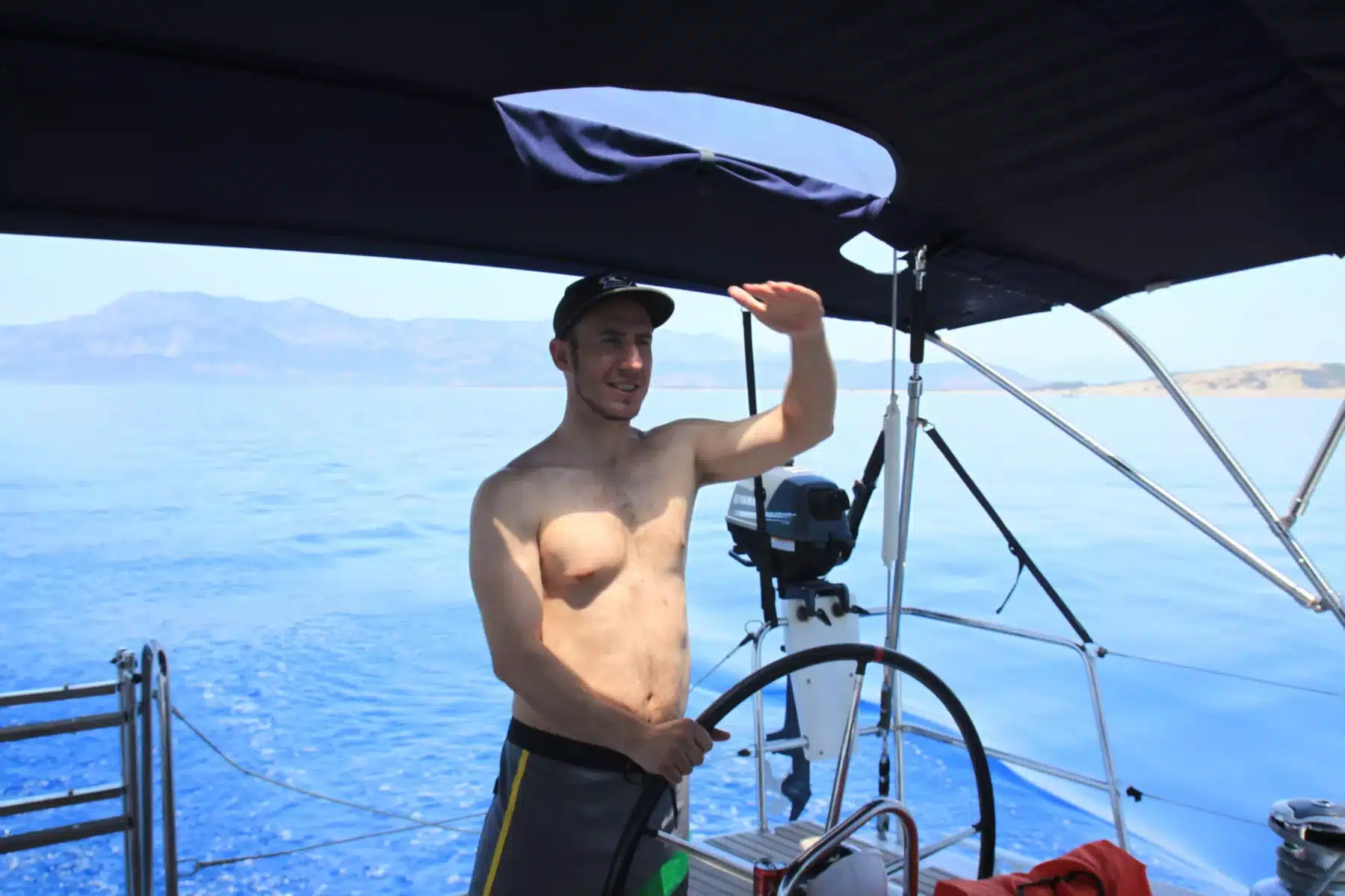 Ein Mann mit Hut und Shorts steht auf einem Katamaran und hält sich an einem Baldachinrahmen fest. Im Hintergrund sind klares blaues Wasser und Berge zu sehen. Er lächelt und scheint einen sonnigen Tag auf dem Wasser zu genießen.