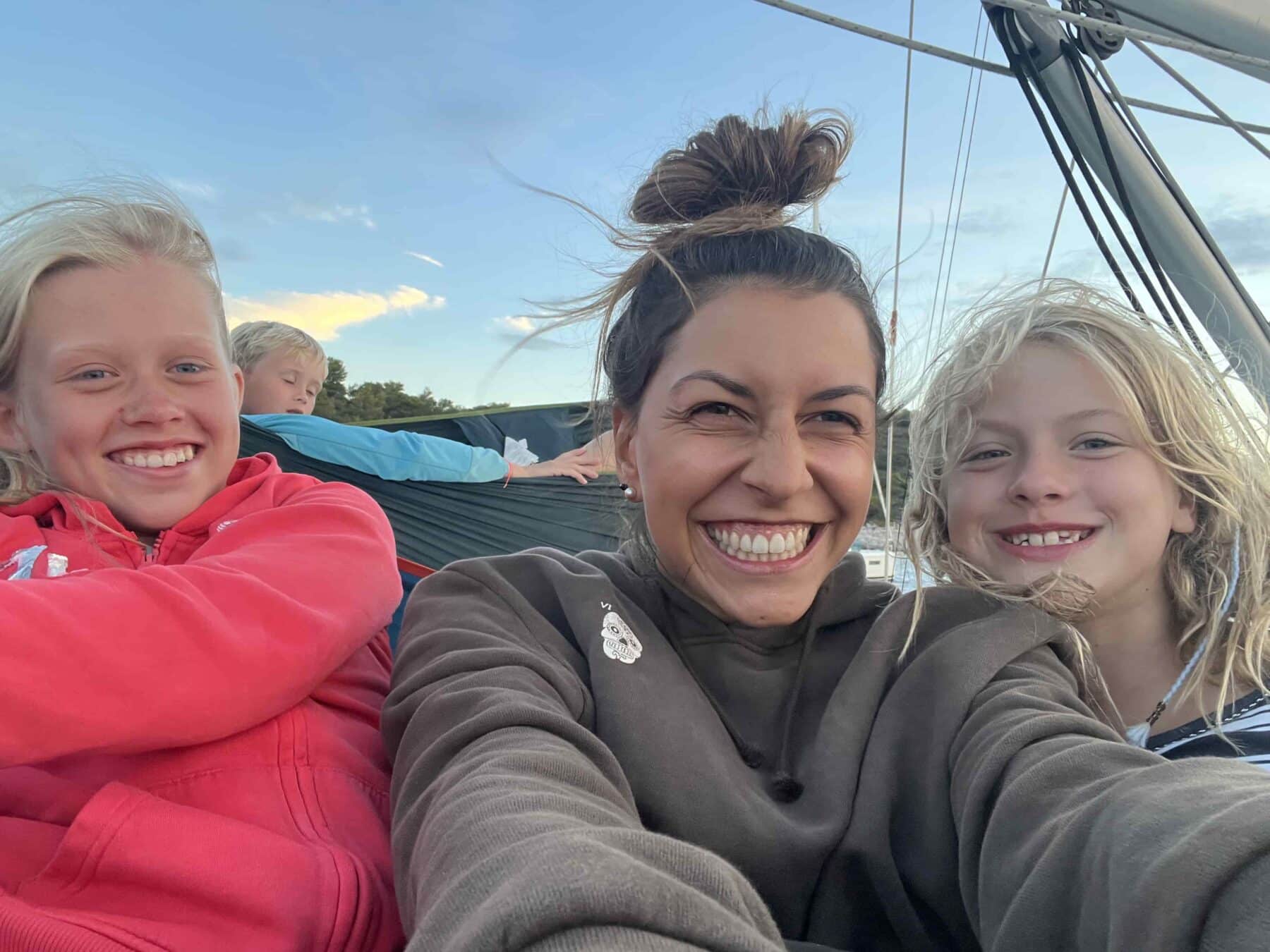 Eine Frau mit Dutt und zwei Kindern sitzt auf einer Segelyacht und lächelt breit. Das Kind links trägt einen roten Kapuzenpullover, das Kind rechts hat lange blonde Haare und trägt einen braunen Kapuzenpullover. Im Hintergrund sind die Struktur des Bootes und der blaue Himmel zu sehen.