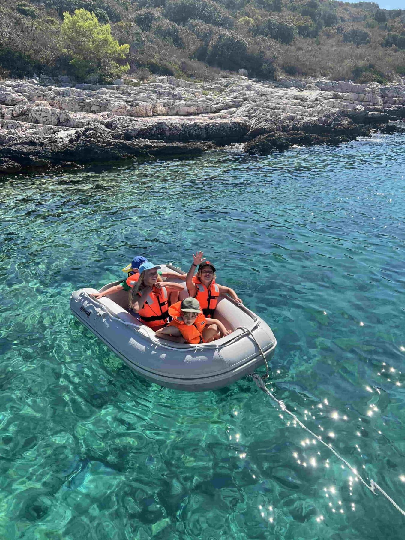 Drei Menschen in orangefarbenen Schwimmwesten sitzen auf einem Katamaran auf klarem, türkisfarbenem Wasser in der Nähe einer felsigen Küste und genießen einen sonnigen Tag.