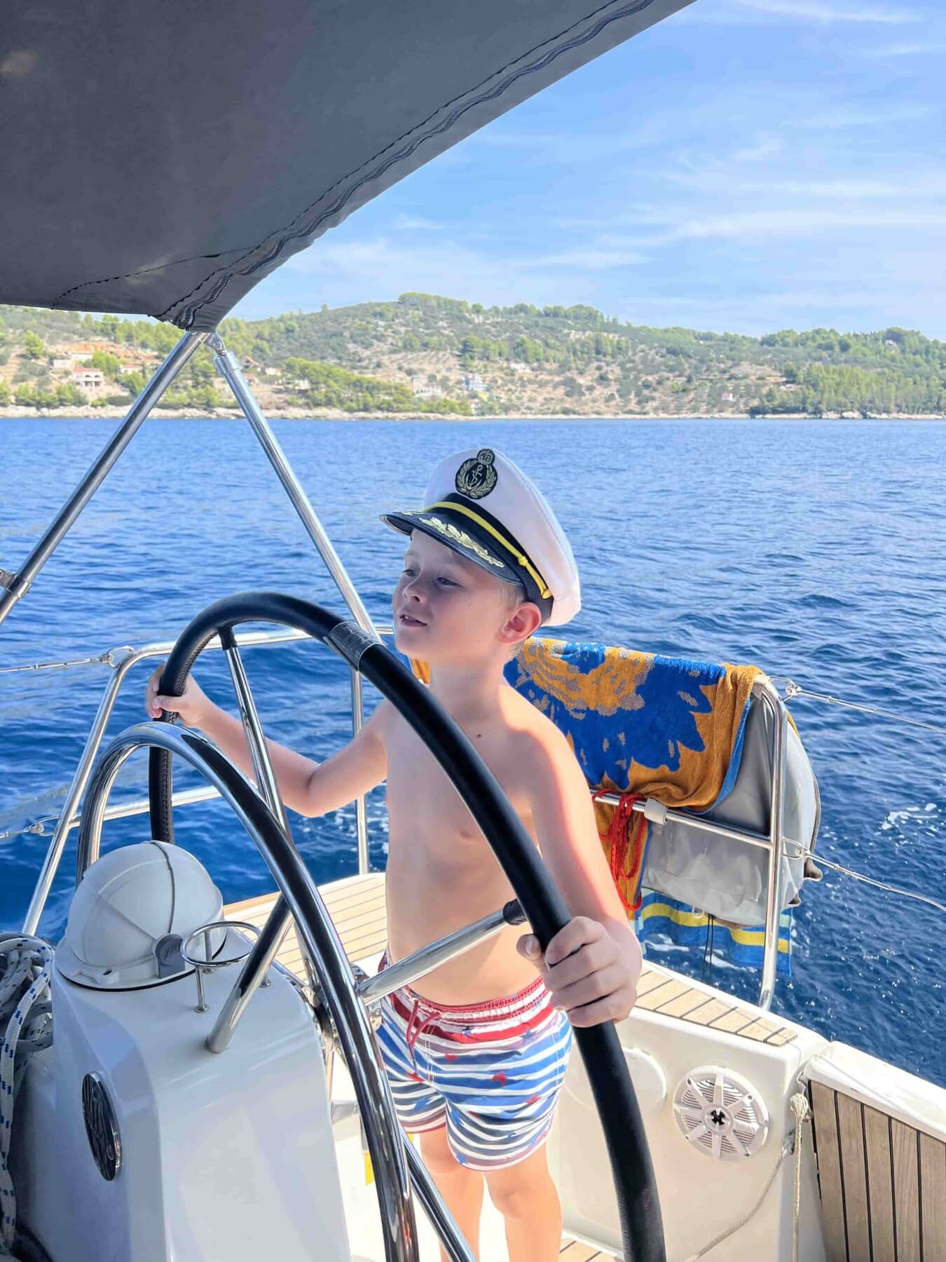 Ein Kind mit weißer Kapitänsmütze, bunter Badehose und ohne Hemd steht selbstbewusst am Steuerrad einer Segelyacht. Das Boot liegt in blauen, ruhigen Gewässern mit Hügeln und Grün im Hintergrund. Ein Handtuch hängt über der Reling des Bootes und fängt die Essenz einer ruhigen Segelreise ein.
