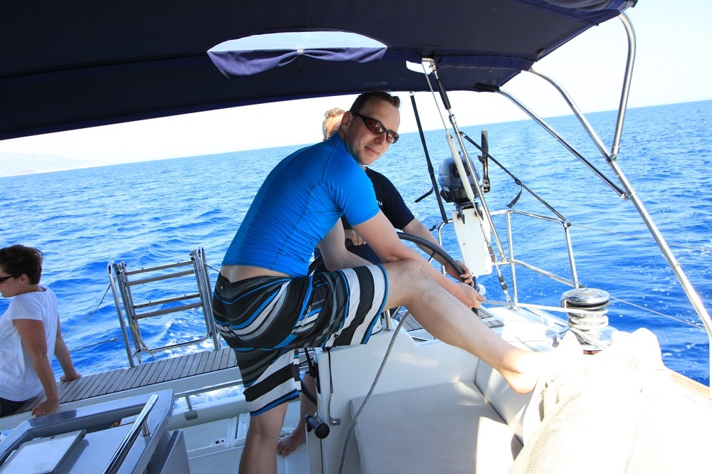 Ein Mann mit Sonnenbrille und blauer Sportkleidung bedient unter klarem Himmel eine Winde auf einem Katamaran. Eine weitere Person ist teilweise im Hintergrund zu sehen. Sie befinden sich auf dem Meer, in der Ferne ist eine Küstenlinie zu sehen.