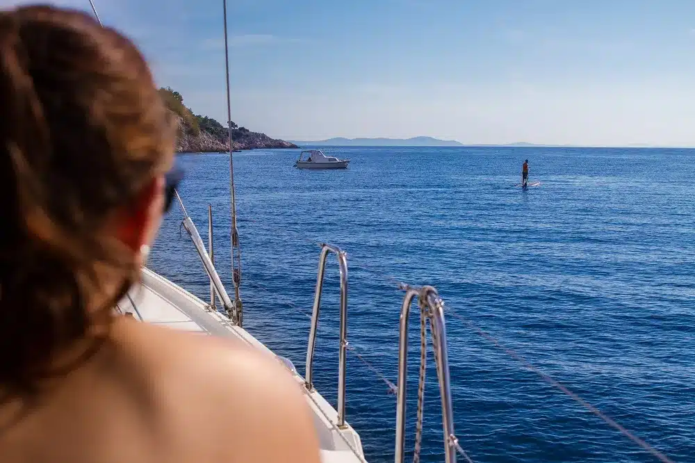 Eine Frau blickt von einer Segelyacht aus auf das ruhige Meer und sieht in der Ferne unter einem klaren blauen Himmel eine Person auf einem Paddleboard und ein weiteres Boot.