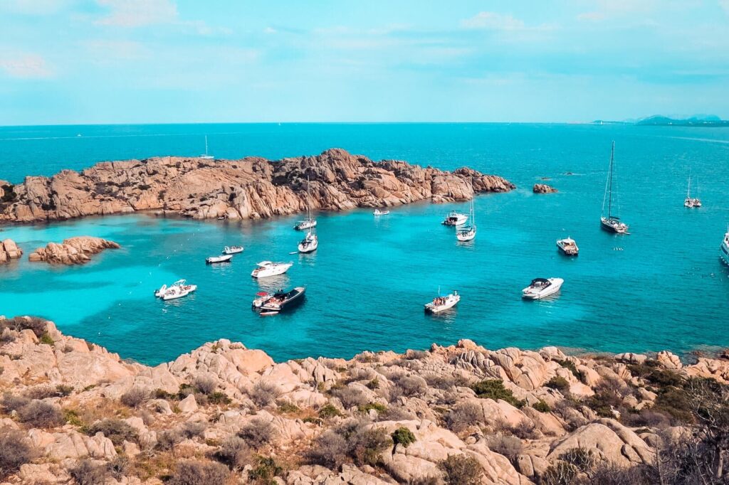 Eine malerische Küstenszene mit mehreren Motorbooten und einer Segelyacht, die in einer ruhigen, strahlend blauen Bucht vor Anker liegt. Die felsige Küste ist mit spärlichem Grün und schroffen Klippen übersät, und das ruhige Meer erstreckt sich unter einem klaren, sonnigen Himmel bis zum fernen Horizont – perfekt für einen Segeltörn.