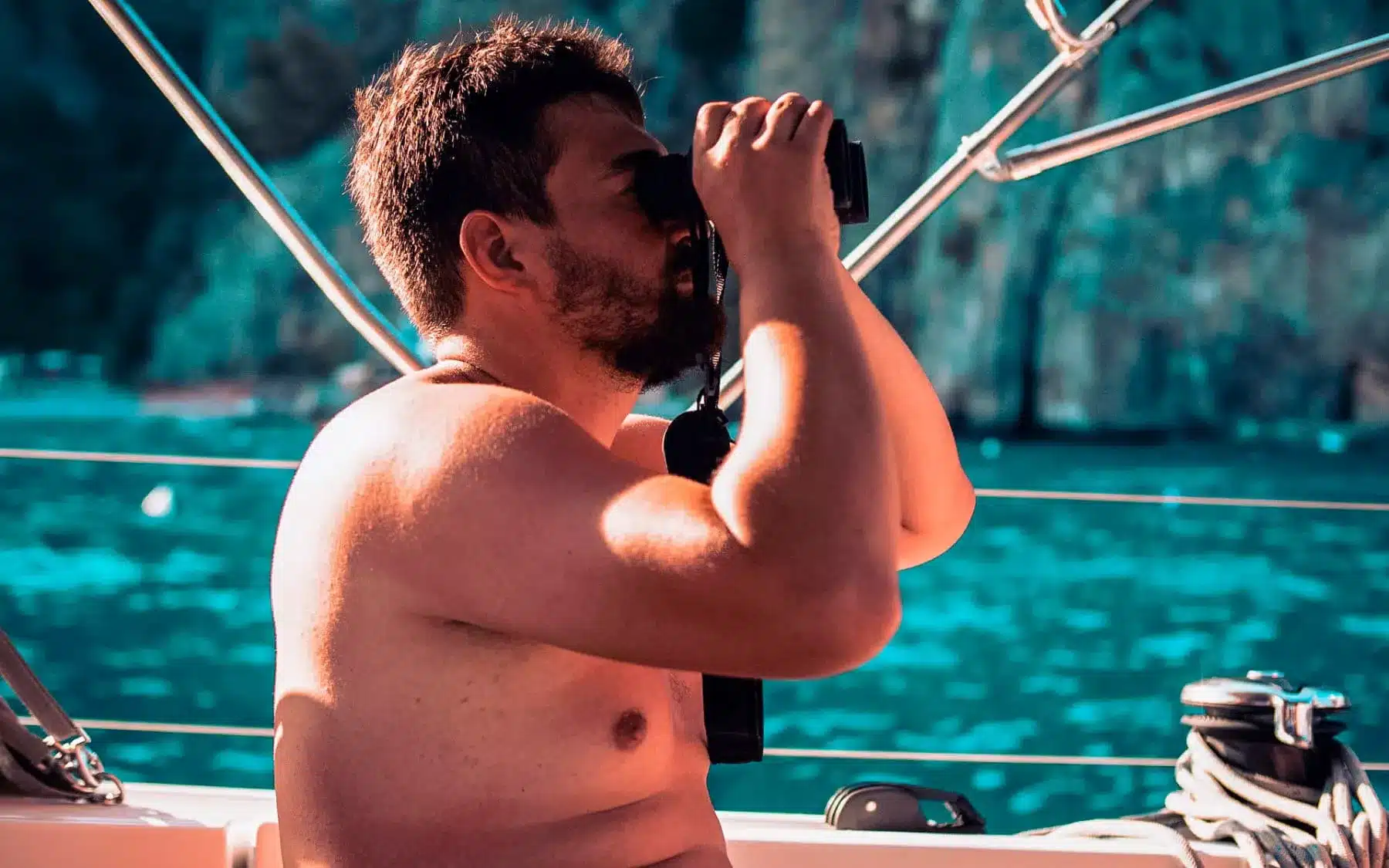 Ein bärtiger Mann ohne Hemd benutzt ein Fernglas auf einer Segelyacht, umgeben von einer ruhigen blauen Wasserkulisse.