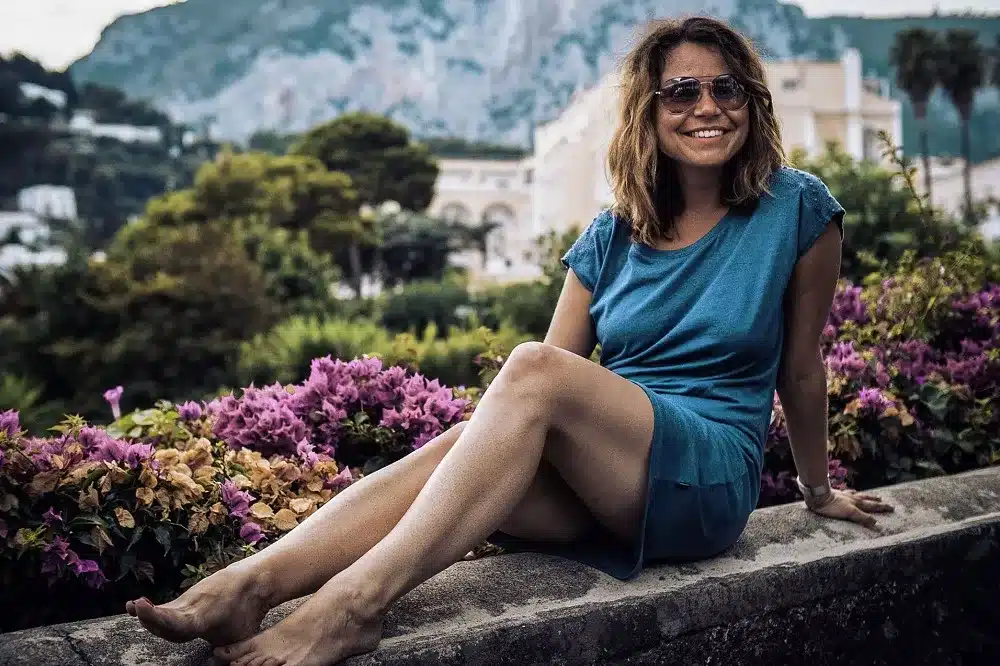 Eine Frau in einem blauen Kleid sitzt während ihres Segelurlaubs barfuß lächelnd auf einem Steinvorsprung, umgeben von üppigem Grün und rosa Blumen, mit einer Bergkulisse im Hintergrund.