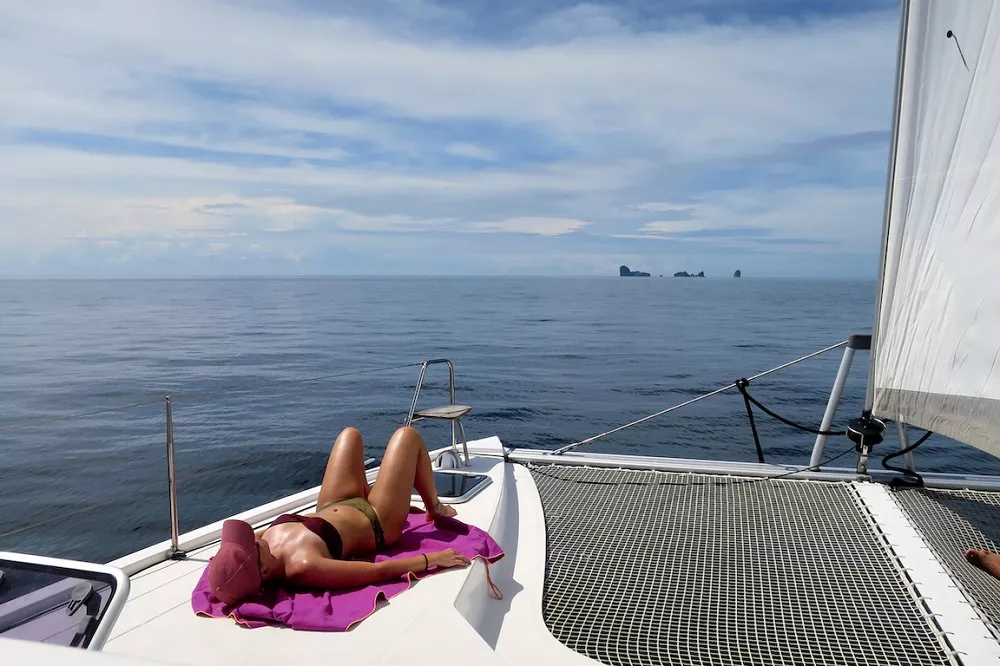 Eine Person liegt auf dem Deck eines Segelboots und sonnt sich vor dem ruhigen Meer und in der Ferne sind Inseln zu sehen. Die weißen Segel des Segelboots sind während des Segeltörns rechts teilweise zu sehen.