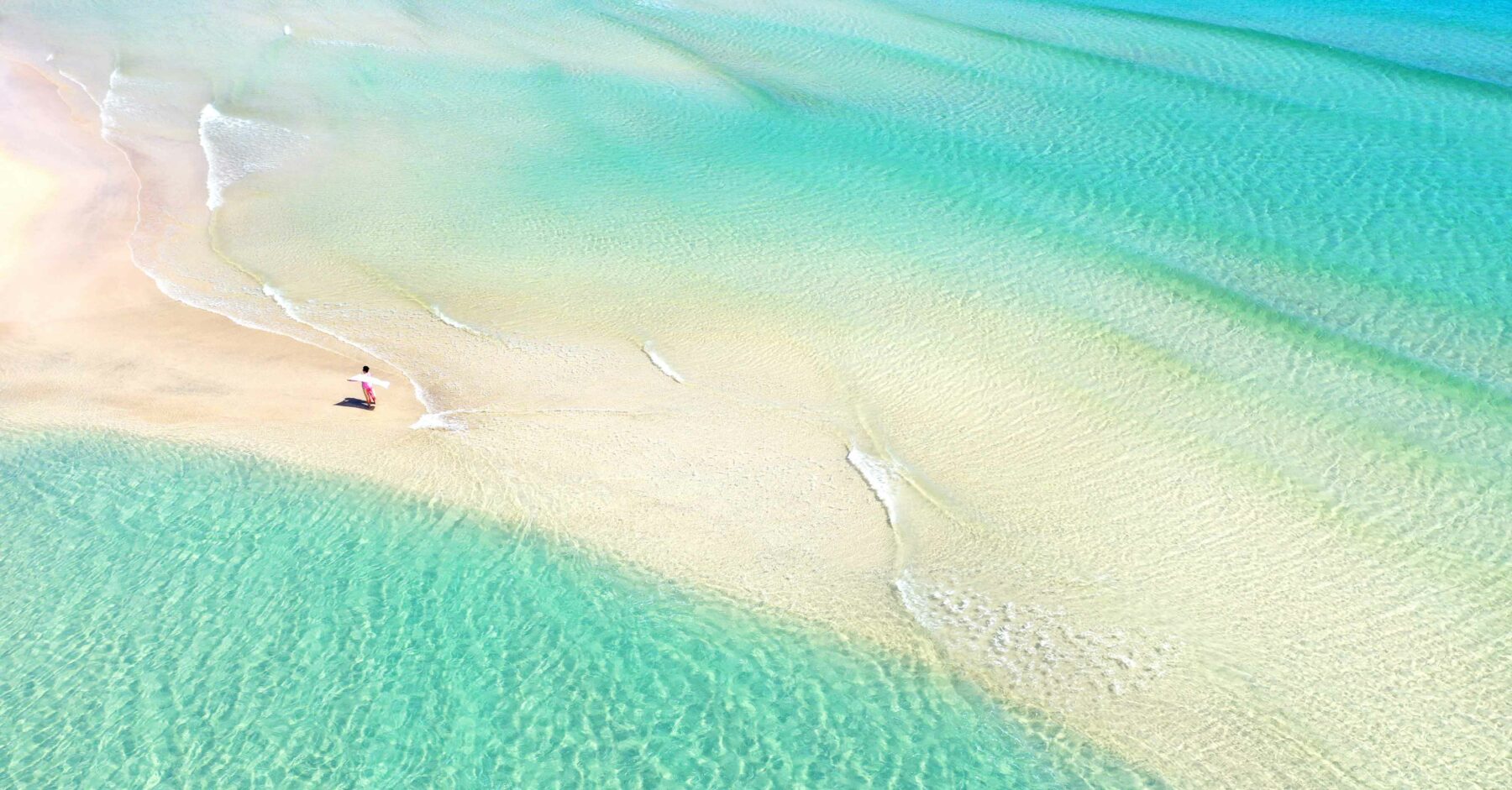 Luftaufnahme einer Person, die an einem unberührten Sandstrand spazieren geht, an dessen Ufer türkisfarbenes Wasser sanft schwappt und die Sonne sanfte Schatten auf eine nahe gelegene Segelyacht wirft.