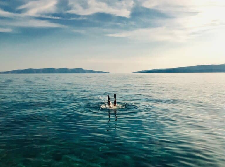Eine Person taucht in das klare, blaue Wasser eines ruhigen Meeres ein, mit entfernten Hügeln und einem Katamaran unter einem klaren Himmel im Hintergrund.