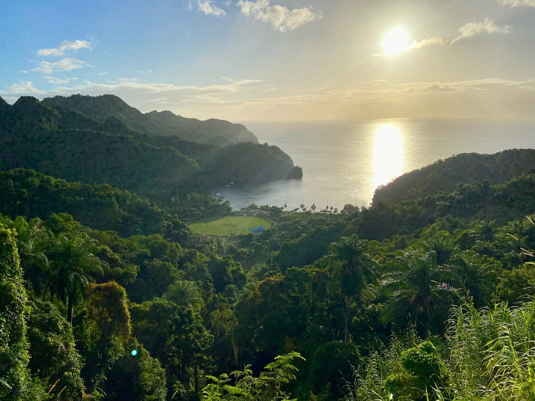 Sonnenuntergang über einer malerischen Küstenlandschaft mit üppigen Hügeln, einer ruhigen Bucht und tropischen Bäumen unter einem strahlenden Himmel während einer Segelreise.
