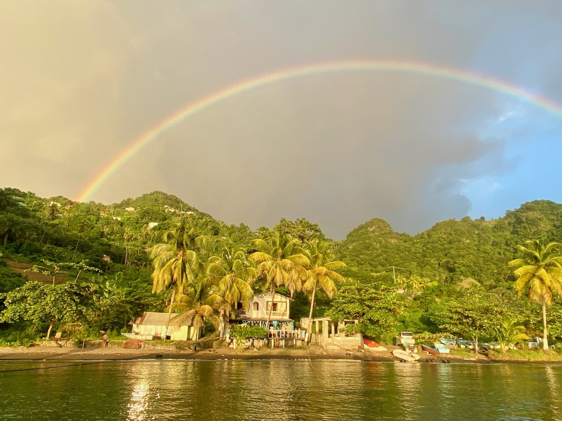 Ein leuchtender Regenbogen, der sich über eine üppige tropische Landschaft mit Palmen und schlichten Gebäuden in der Nähe eines ruhigen Flusses spannt, unter einem teilweise bewölkten Himmel bei Sonnenuntergang, schafft die Kulisse für einen idyllischen Segelurlaub.