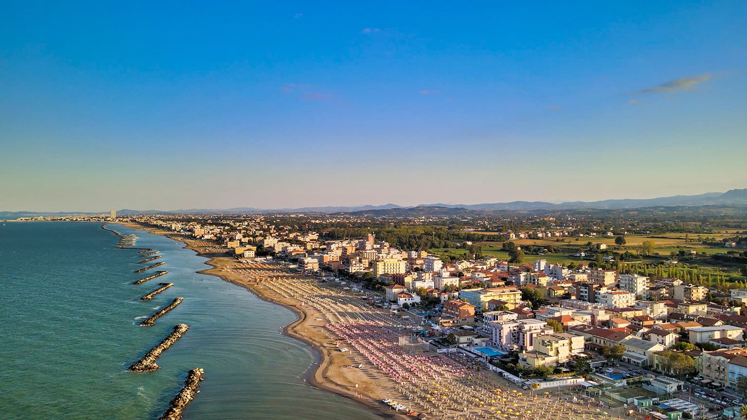Luftaufnahme einer Küstenstadt bei Sonnenuntergang. Zu sehen ist ein langer Strand mit bunten Sonnenschirmen und eine dicht besiedelte, sich in die Ferne erstreckende, von Bergen begrenzte Stadt, ideal für einen Segelurlaub.
