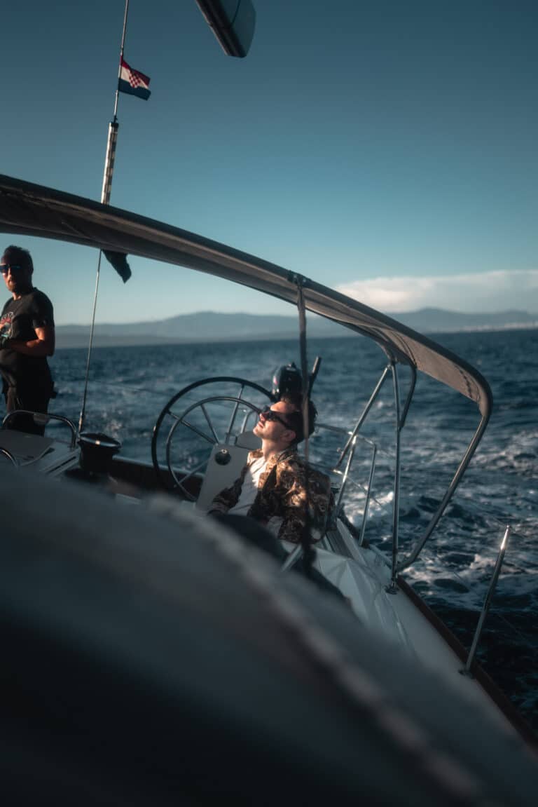 Eine Frau steuert eine Segelyacht und blickt auf die Segel, im Hintergrund das Meer und den blauen Himmel. Eine andere Person steht in der Nähe und auf dem Boot weht eine amerikanische Flagge.