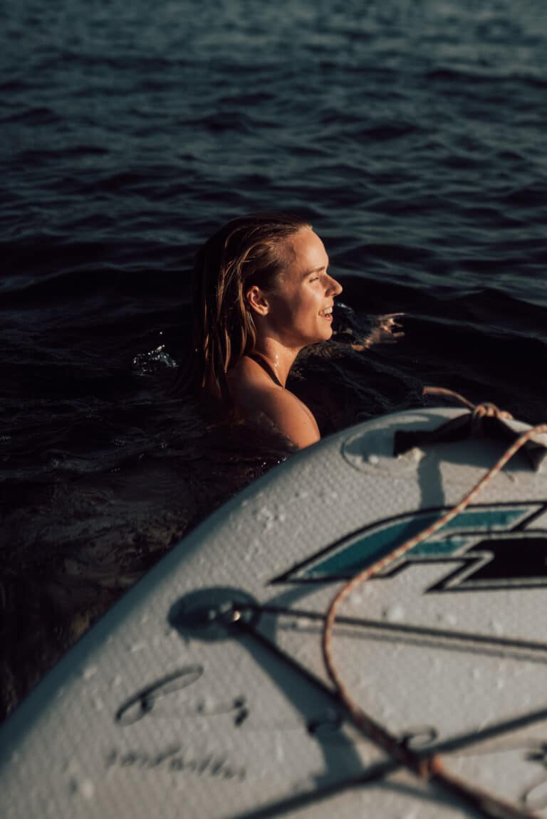 Eine Frau mit ihrem Surfbrett genießt das Wasser, lächelt, die Sonne beleuchtet ihr Gesicht und blickt zum Horizont in der Nähe eines Katamarans.