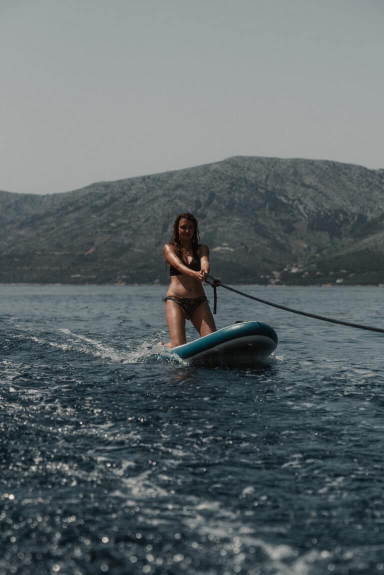 Eine Frau im Bikini macht während ihres Segelurlaubs auf dem Kneeboard eine ruhige See mit Bergen im Hintergrund und nutzt ein Schleppseil, um das Gleichgewicht zu halten.