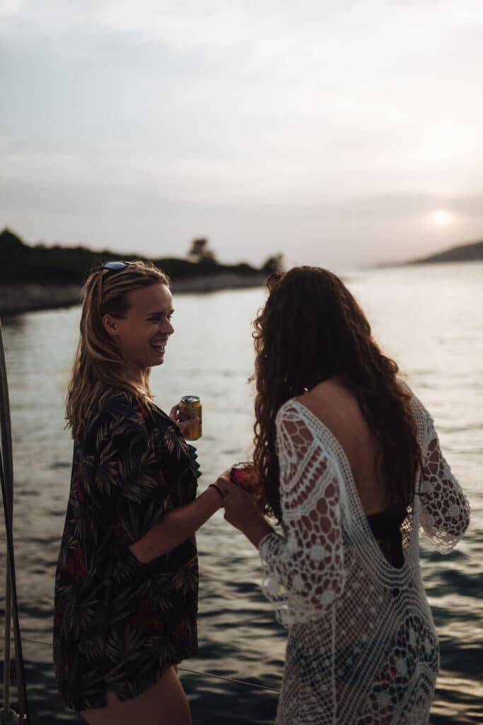 Zwei Frauen genießen Getränke, während sie bei Sonnenuntergang neben einem ruhigen Gewässer stehen. Eine blickt lachend in die Kamera und hält eine Dose, während die andere mit dem Rücken zur Kamera steht und ein Häkelkleid trägt. Im Hintergrund sind Bäume und fernes Land zu sehen, was auf einen Nachmittag voller Mitsegeln-Abenteuer hindeutet.