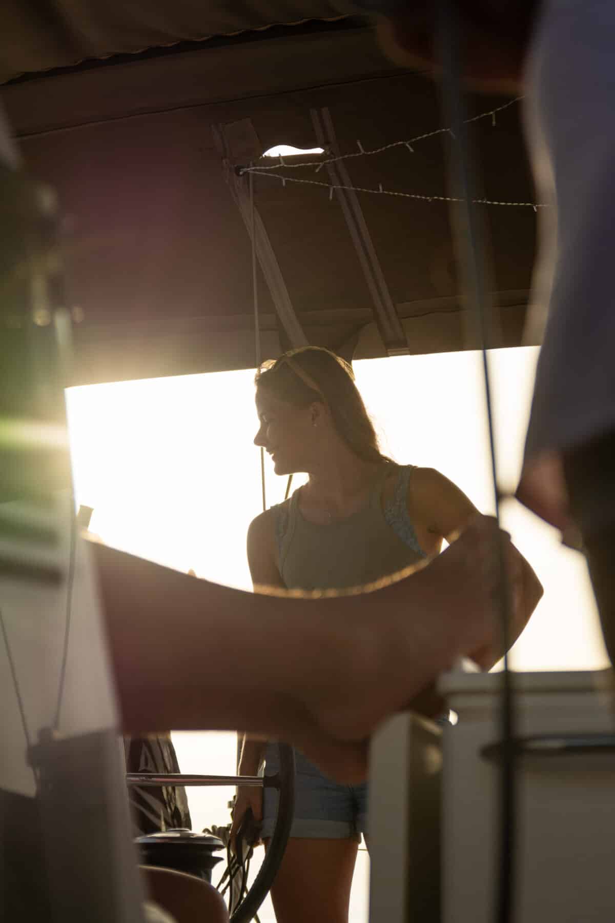 Frau steht auf einer Segelyacht, im Gegenlicht der Sonne, und unterhält sich mit einer Person, deren Arm im Vordergrund zu sehen ist. Die Umgebung ist ruhig und strahlt warmes Licht aus.