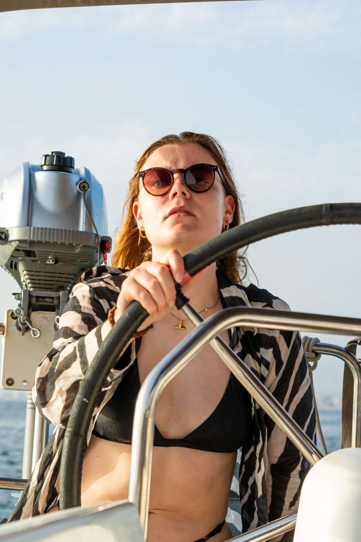 Eine Person mit langen Haaren, Sonnenbrille und schwarzem Bikini steuert eine Segelyacht. Sie hat einen konzentrierten Gesichtsausdruck und hält das Steuer während ihres Segelurlaubs mit beiden Händen. Der Hintergrund zeigt einen blauen Himmel und Wasser an einem sonnigen Tag.