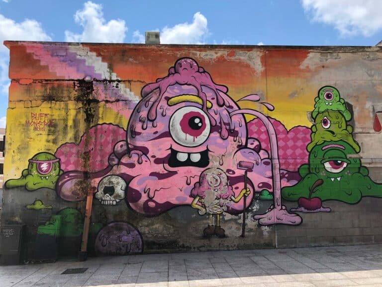 Ein farbenfrohes Street-Art-Wandbild zeigt in der Mitte eine große rosafarbene, einäugige Kreatur, von der Schleim tropft und die eine Farbrolle hält. Um sie herum sind weitere grüne und violette Monster, ein Totenkopf und abstrakte Formen zu sehen. Der helle Himmel darüber spiegelt einen lebhaften Katamaran-Segelurlaub mit vereinzelten Wolken wider.