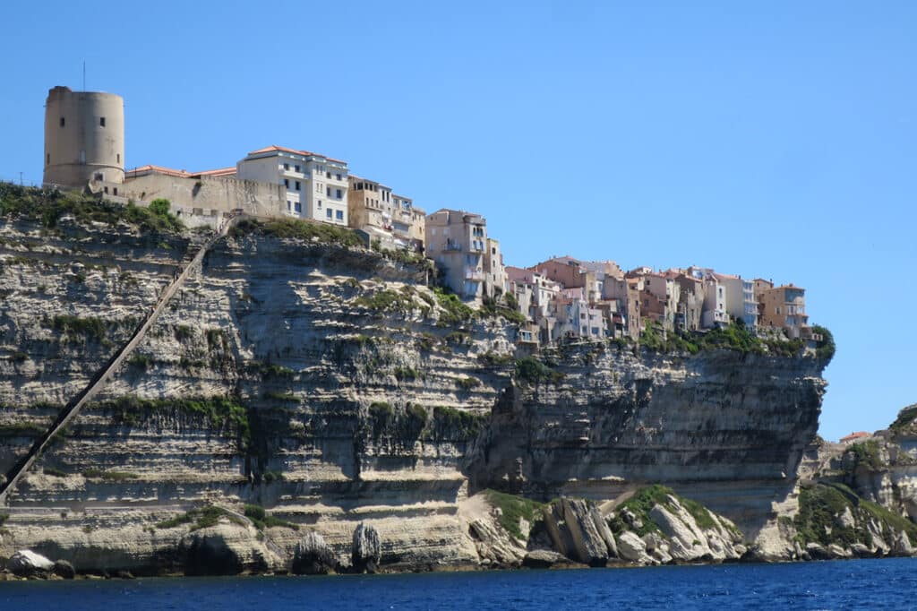 Küstenlandschaft mit einer Klippe, auf der sich eine Reihe pastellfarbener Gebäude und ein alter Turm befinden, unter einem klaren blauen Himmel und darunter dem Meer, auf dem Katamarane kreuzen.