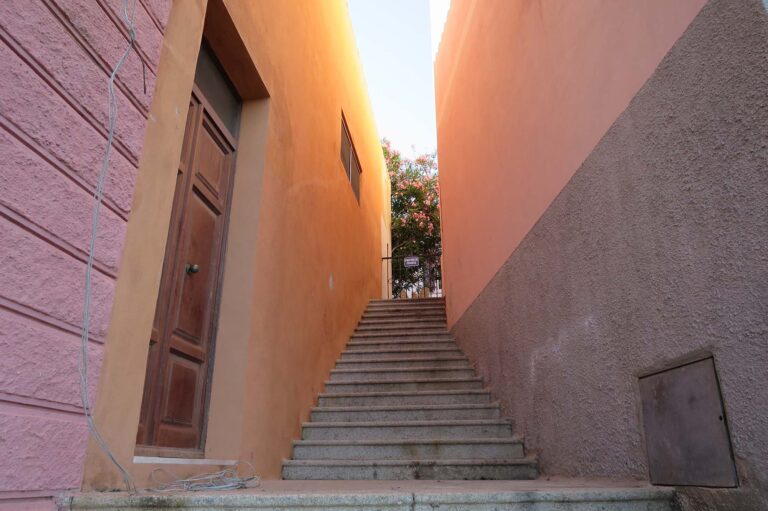 Schmaler Pfad zwischen zwei pfirsichfarbenen Gebäuden mit einer Betontreppe, die nach oben zu einem blühenden Baum führt, der unter einem klaren Himmel an der Spitze zu sehen ist.