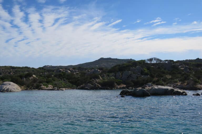 Eine heitere Meereslandschaft mit ruhigem blauen Wasser im Vordergrund, felsigen Küstenlinien mit viel Grün und hügeligem Gelände im Hintergrund unter einem von dünnen Wolken durchzogenen Himmel, perfekt für eine Segelreise.