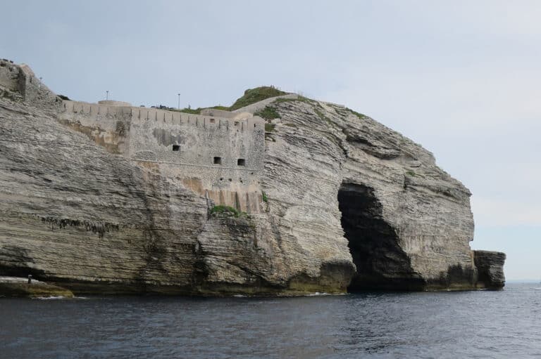 Eine Festung thront auf einer schroffen Klippe mit horizontalen Rillen und bietet Blick auf ein ruhiges Meer. Die Klippe hat eine große dunkle Öffnung, möglicherweise eine Meereshöhle. Der Himmel ist hellblau mit ein paar vereinzelten Wolken, perfektes Wetter zum Mitsegeln während Ihrer Segelreise.