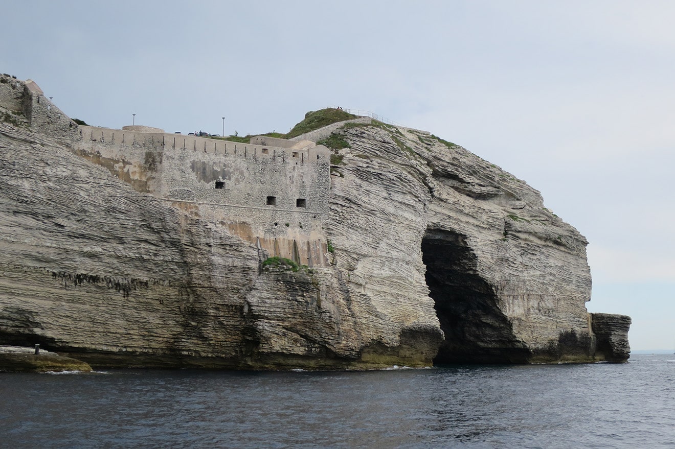 Eine Festung thront auf einer schroffen Klippe mit horizontalen Rillen und bietet Blick auf ein ruhiges Meer. Die Klippe hat eine große dunkle Öffnung, möglicherweise eine Meereshöhle. Der Himmel ist hellblau mit ein paar vereinzelten Wolken, perfektes Wetter zum Mitsegeln während Ihrer Segelreise.