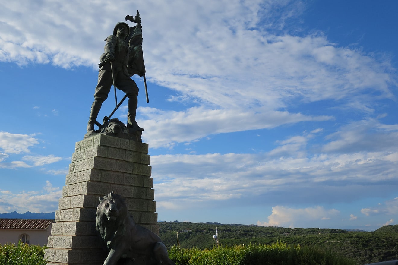 Auf einem Steinsockel steht die Statue eines Mannes, der eine Flagge hält. Er wirkt siegreich, mit einem Fuß auf einem Helm, was an die Triumphe erinnert, die man während eines abenteuerlichen Segelurlaubs erlebt. Am Fuß des Sockels steht ein Löwe. Der Hintergrund zeigt einen strahlend blauen Himmel mit vereinzelten Wolken und grünen Hügeln in der Ferne.