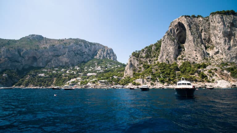 Eine malerische Küstenszene zeigt schroffe, mit Grün bedeckte Klippen, blaues Meerwasser und mehrere festgemachte Boote, darunter eine elegante Segelyacht, unter einem klaren Himmel. Die Landschaft ist übersät mit zahlreichen kleinen Bauten, die zwischen den Klippen eingebettet sind.