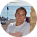 Eine Frau in einem weißen T-Shirt hält eine große Eistüte und steht im hellen Sonnenlicht am Meer mit einem klaren blauen Himmel und einem Katamaran im Hintergrund.