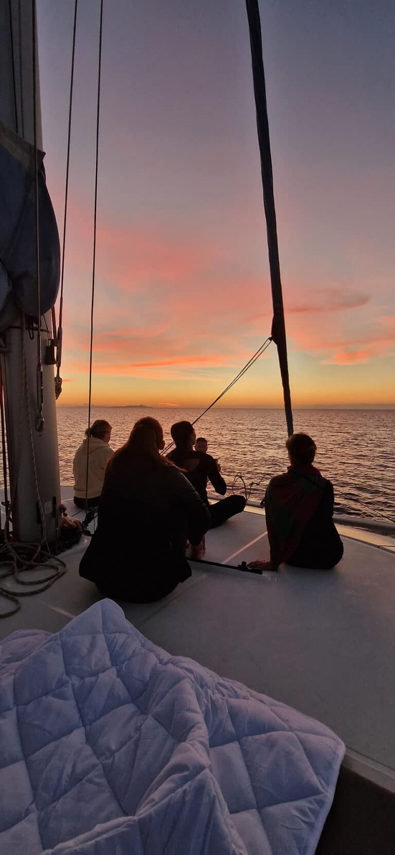 Vier Personen sitzen bei Sonnenuntergang auf dem Deck einer Segelyacht und blicken auf einen leuchtend orange-rosa Himmel über einem ruhigen Meer.