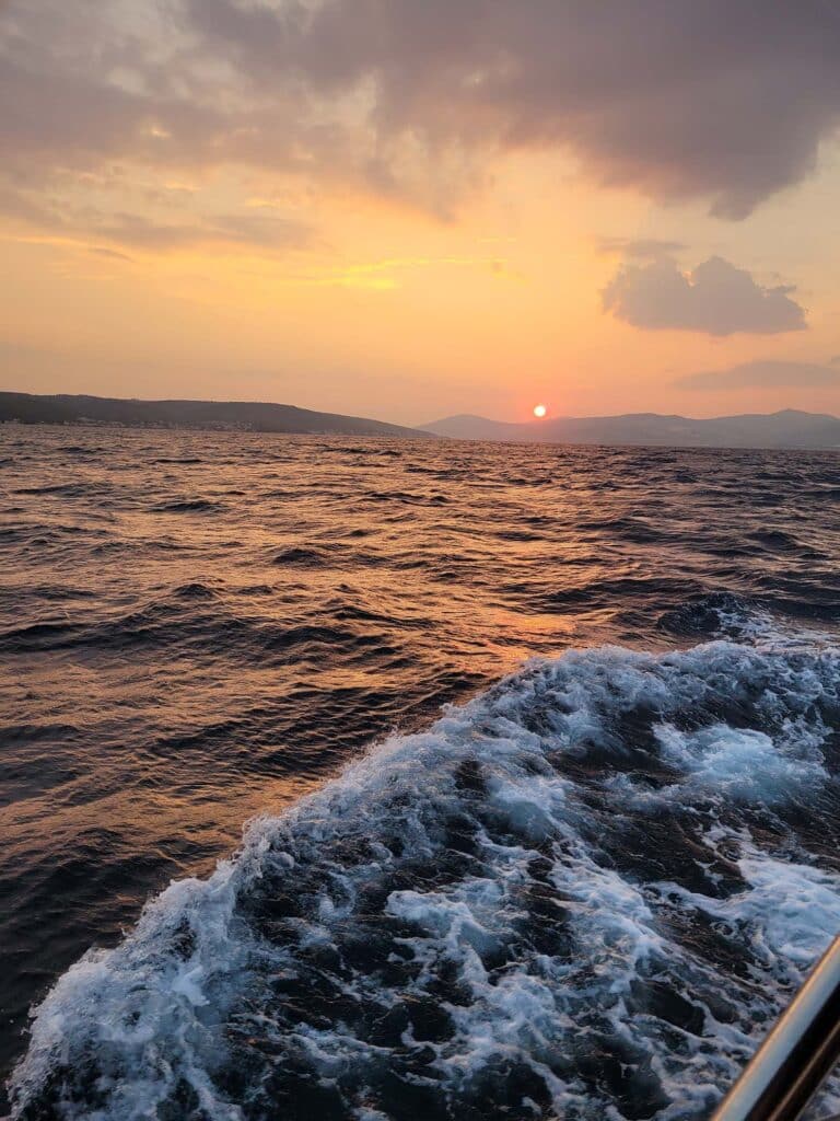 Ein malerischer Blick auf einen Sonnenuntergang über dem Meer, mit sanften Wellen, die einen Kräuseleffekt auf der Wasseroberfläche erzeugen, perfekt für einen privaten Segelurlaub. Der Himmel ist in Orange-, Gelb- und Rosatönen gemalt und in der Ferne sind Hügel am Horizont zu sehen.