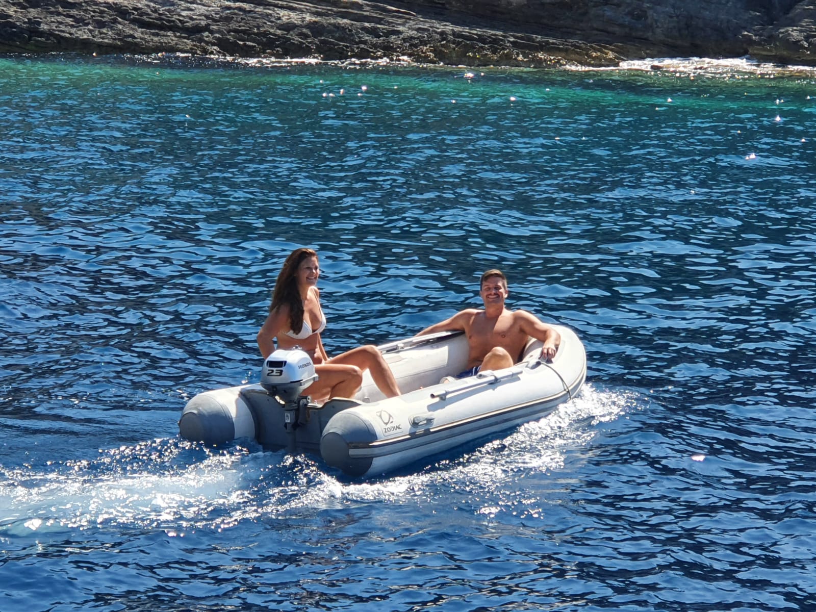 Zwei Personen genießen einen sonnigen Tag auf einem kleinen Katamaran im klaren blauen Wasser nahe einer felsigen Küste.